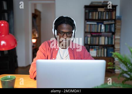 Fokussierter junger schwarzer Fernarbeiter, der Kopfhörer trägt und Musik hört, während er von zu Hause aus arbeitet Stockfoto