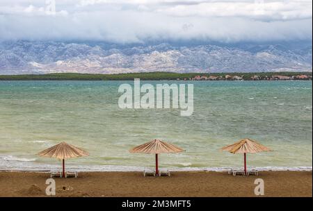 Nin, Kroatien - drei Schilf-Sonnenschirme am leeren Queen's Beach bei der Stadt Nin am Ende des Sommers mit Velebit-Bergen im Hintergrund und türkisfarbenem Hintergrund Stockfoto