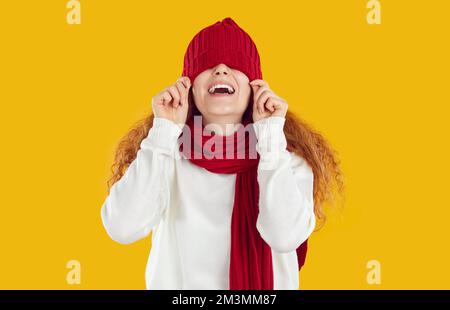 Lachendes glückliches Mädchen mit rotem Strickschal und Hut, die über die Augen auf gelbem Hintergrund gezogen wurden. Stockfoto