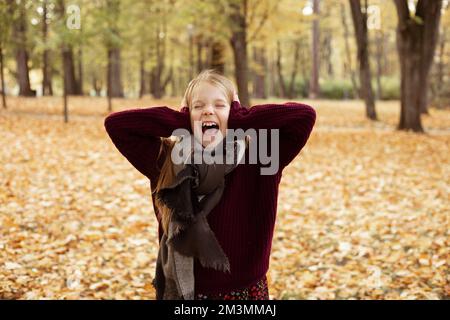 Porträt eines verrückten Teenagers, das im Park zwischen gelben Ahornblättern steht, schreit, Ohren mit Händen bedeckt. Stockfoto