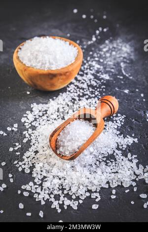 Reines natürliches Meersalz in Holzschüssel und Holzlöffel auf schwarzem Hintergrund Stockfoto