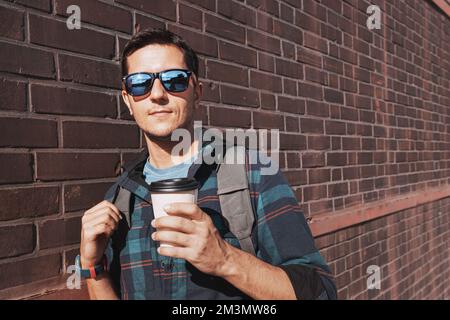 Ein stilvoller junger Mann mit einer Tasse Kaffee zum Mitnehmen posiert vor dem Hintergrund einer orangefarbenen Backsteinwand in der Fabrik Stockfoto