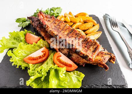 Eine große Sorte Schweinerippchen auf einem Schieferbrett mit Salat, Tomatenscheiben und gegrillten Kartoffeln. Das Konzept der im Garten gekochten Speisen Stockfoto