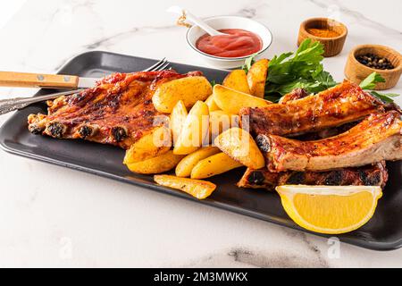 Köstliche Schweinerippchen und gegrillte Kartoffelscheiben, serviert auf einem rechteckigen Keramikgericht mit Petersilie. Das Konzept des Servierens der Speisen. Menü Stockfoto