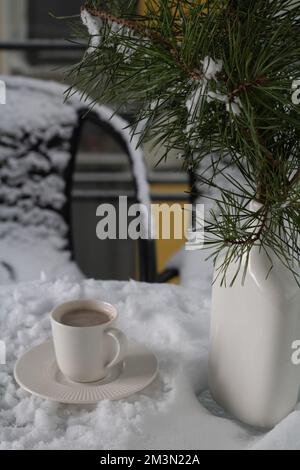 Fotografieren Sie eine weiße Tasse Kaffee auf einer Untertasse neben einer Vase mit Tannenzweigen auf einem mit Schnee bedeckten Tisch Stockfoto