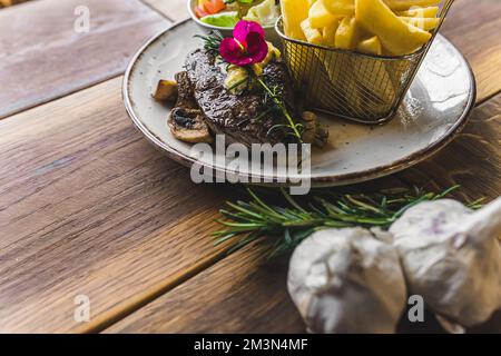 Leckeres Striploin-Steak auf Pilzen mit Pommes frites. Knoblauch im Vordergrund. Holztisch als Hintergrund. Hochwertiges Foto Stockfoto