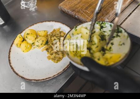 Koch verwendet Kochzangen, um gekochte Kartoffeln neben traditionellem geschmorten Kohl auf einen weißen Teller zu legen. Polnische Küche in einem Restaurant. Hochwertiges Foto Stockfoto