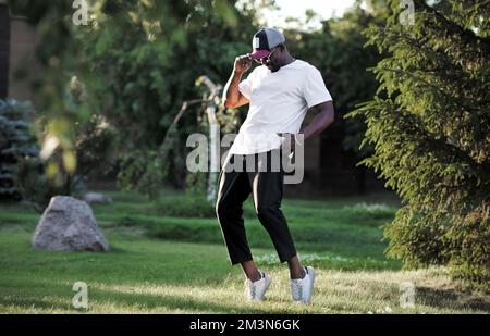 Ein afroamerikanischer Mann tanzt draußen im Naturpark. Tänzerin trägt weißes T-Shirt, schwarze Hose und Baseballkappe Stockfoto