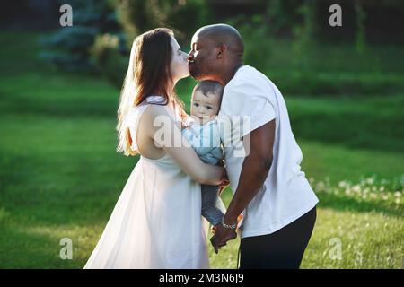 Multiethnische Familie. Kaukasische Mutter und afroamerikanischer Vater küssen und halten Kleinkind in den Armen. Eltern, Porträt von Mom, Dad und Baby auf der Hand Stockfoto