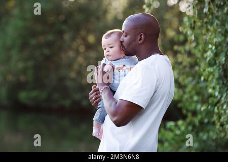 Vater und Sohn. Der afroamerikanische Vater küsst, hält sich an den Armen und liebt ein multiethnisches Kind vor dem Hintergrund der grünen Natur Stockfoto