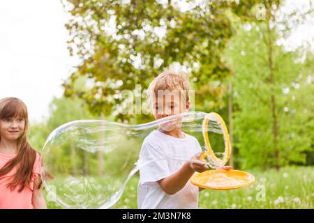 Ein Junge, der eine große Seifenblase durch den Zauberstab wirft, steht neben einer Freundin im Park Stockfoto