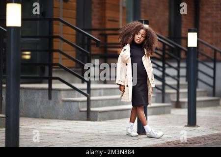 Ein nachdenkliches kleines Mädchen, das auf der Straße steht und auf jemanden wartet, wegschaut, modisch gekleidet in Trenchcoat und schwarzem Kleid. Volle Länge po Stockfoto