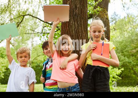 Multirassische Jungen und Mädchen mit Büchern, während sie in den Sommerferien im Park gegen einen Baum stehen Stockfoto