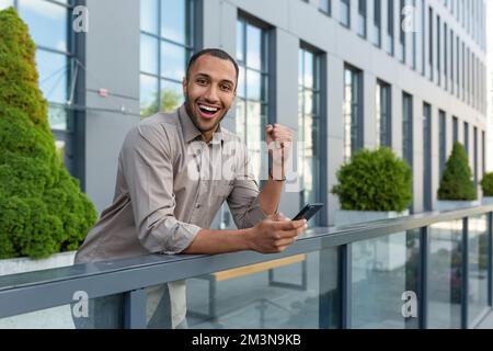 Porträt eines erfolgreichen Geschäftsmanns außerhalb des Bürogebäudes, eines lateinamerikanischen Mannes, der lächelt und vor die Kamera schaut, um den Sieg und die erfolgreiche Leistung zu feiern, eines Mannes, der sein Telefon in der Hand hält und eine Triumph-Geste hochhält Stockfoto