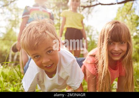 Porträt von glücklichen Kindern, die sich während des Schubkarren-Rennens im Park an sonnigen Tagen gegenseitig gegeneinander antreten Stockfoto