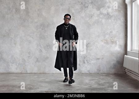 Ein stilvoller Mann, der an die graue Studiowand geht, einen schwarzen Mantel, Rollkragenpullover, Hosen und Slipper trägt. Mode-afroamerikanisches männliches Modell in Fashi Stockfoto