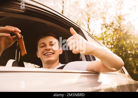 Anfänger halten Autoschlüssel aus dem Fenster, zeigen Daumen hoch positive Geste und lächeln. Konzept für einen neuen Fahrer auf der Straße. Stockfoto