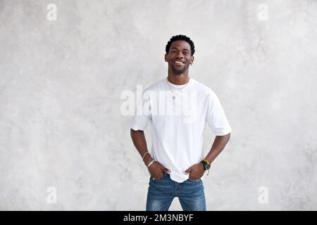 Porträt eines lächelnden jungen afroamerikanischen Mannes gegen eine hell strukturierte Wand im Studio. Das glückliche männliche Model hat die Hände in den Taschen und trägt lässige Kleidung Stockfoto