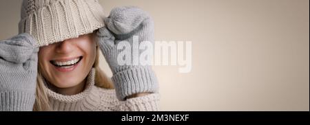 Lächelnde Frau, die warme Kleidung trägt und das Gesicht unter einer Strickmütze aus Wolle auf beigefarbenem Hintergrund verdeckt. Trendige Winterbekleidung. Banner mit Kopierbereich Stockfoto