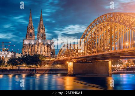 Malerischer Blick auf die beleuchtete Brücke über den Rhein, an der Züge und Touristen vorbeifahren, und den Kölner Dom am Abend zur blauen Stunde. Stockfoto