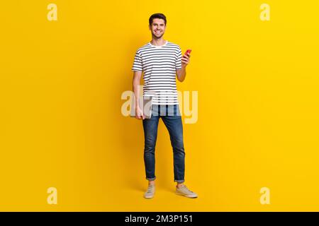 Langes Foto eines coolen, hübschen Mannes, gekleidet mit gestreiftem T-Shirt, funktionierendes Apfel, samsung Modern, isolierter gelber Hintergrund Stockfoto