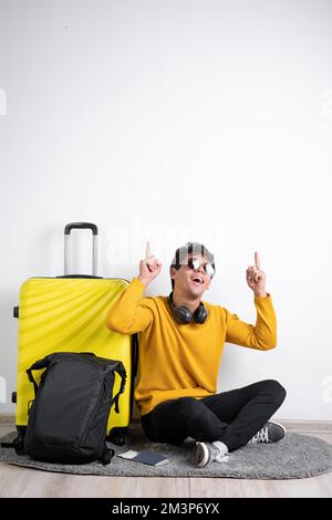 Reisender Tourist Mann in legerem Pullover mit gelbem Koffer sitzen mit den Fingern über dem Bereich isoliert auf weißem Hintergrund. Flugflugkonzept Stockfoto