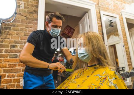 Männlicher Friseur mit Maske, der die blonden Haare seiner Klientin kämmt, im professionellen Friseursalon. Mittlere Aufnahme. Hochwertiges Foto Stockfoto