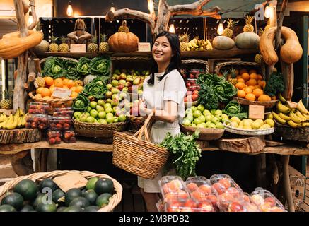 Seitenansicht einer asiatischen Frau mit Korb-Warenkorb auf einem lokalen Markt. Lächelnde Frau, die Bio-Lebensmittel kauft. Stockfoto