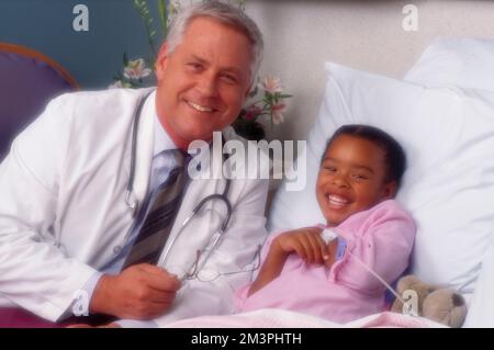 Ein älterer männlicher Arzt, der am Rande des Bettes einer afroamerikanischen Patientin saß, beide lächelten in die Kamera Stockfoto