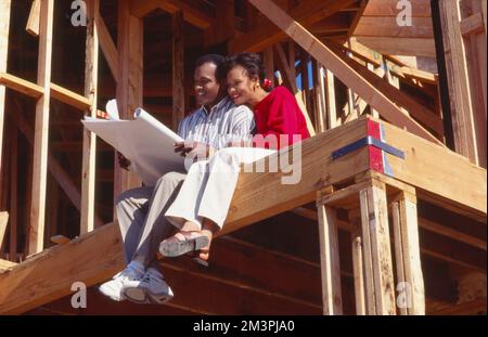 Ein jüngeres schwarzes Paar sitzt im neuen Haus im Bau und schaut sich blaue Abdrücke an Stockfoto