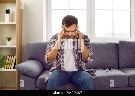 Der junge Mann, der sich die Schläfen reibt, leidet an Migräne, während er zu Hause auf der Couch sitzt. Stockfoto