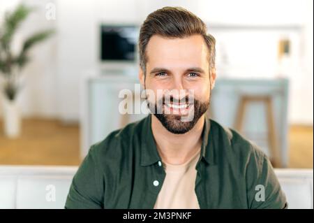 Foto eines hübschen, charismatischen, attraktiven, weißen Bärtigen Mannes, der ein elegantes, legeres Hemd trägt, zu Hause sitzt, in die Kamera schaut, freundlich und glücklich lächelt Stockfoto