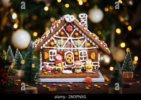 Weihnachtshaus aus Lebkuchen. Verspielt dekoriert mit bunten Gummibären. Weihnachtsbaum im Hintergrund mit Lichtern. Festliches Neujahrsatmo Stockfoto