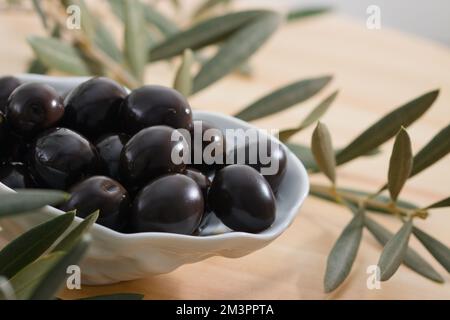 Detail einer Keramikschüssel mit schwarzen Oliven neben einigen Olivenzweigen auf einem Holztisch Stockfoto