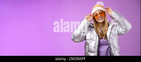 Stilvolle freche gewagte blonde europäische Mädchen handeln cool tragen stilvoll Sonnenbrille Silber Jacke Winter Hut Überprüfung Rahmen Nase kippbaren Kopf Frech lächelnd Stockfoto