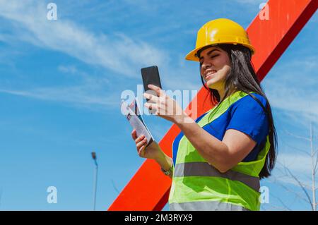 Die junge, weiße Latina-Ingenieurin, eine Arbeiterin auf einer Baustelle, lächelt gerne und überprüft ihr Handy draußen, Kopierraum. Stockfoto