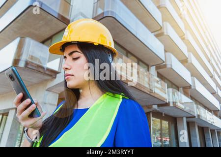 Nahaufnahme einer weißen jungen Frau, Ingenieur, der an einem Gebäude steht, ihr Handy schickt, den Bildschirm liest, Industrie und Technologie C Stockfoto