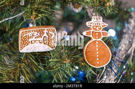 Eine kleine Portion süßen Lebkuchen-Gebäckstücks, das an einem prunkvollen Weihnachtsbaum hängt. Gebackene Weihnachtskekse mit Winterstimmung in Schneemannform oder bemalte Kirche. Stockfoto