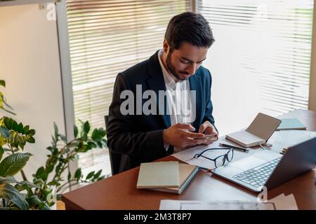 Ein gutaussehender indischer Geschäftsmann im Anzug benutzt sein Telefon, wenn er vom Heimbüro aus arbeitet Stockfoto