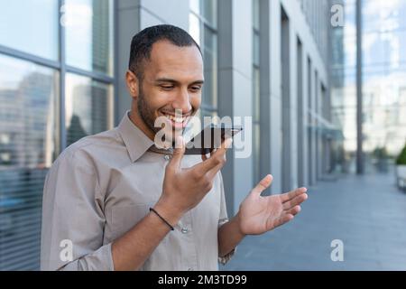 Lächelnder hispanischer Geschäftsmann, der Sprachnachrichten aufnimmt, glücklicher Mann außerhalb des Bürogebäudes, der Befehle auf dem Smartphone für die KI-Suche liest. Stockfoto