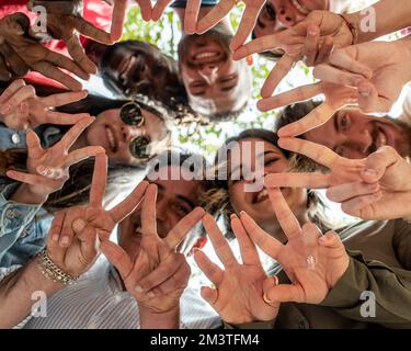 Gruppe glücklicher Freunde, die im Park im Kreis stehen und ein Zeichen für Frieden oder Sieg zeigen, Blick von unten, sieben Personen aus einer multirassischen Familie Stockfoto