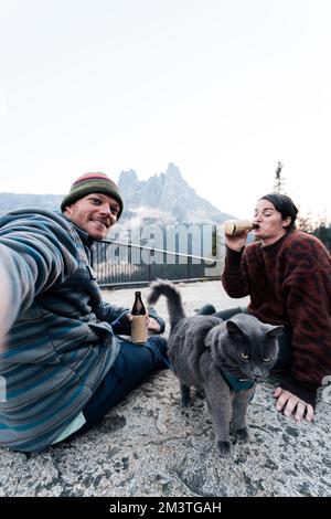 Die junge 30s-jährige weiße Frau sitzt mit ihrem Mann und ihrer Katze und genießt einen Drink, während sie ihre graue russische blaue Katze an der Leine im Wildnis-Wald hält Stockfoto