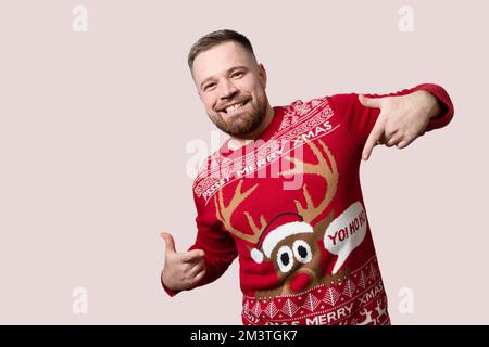Junger, bärtiger Mann, der seinen Rentier-Weihnachts-Pullover zeigt, isoliert auf rotem Hintergrund. Speicherplatz kopieren Stockfoto