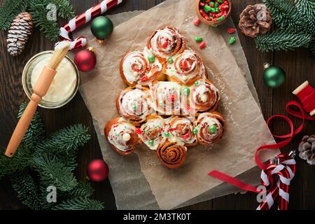 Weihnachtlich baumförmige Zimtrollen oder Zimtbrötchen mit Zimt und Sahne-Sauce auf Holzhintergrund. Festliche Idee für hausgemachten, traditionellen Winter Stockfoto