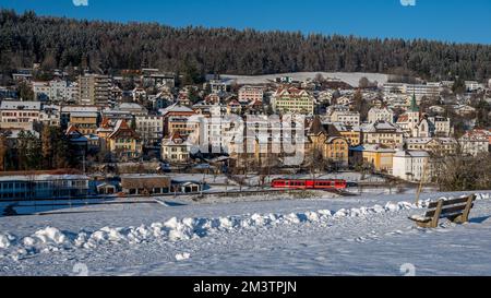 Winterlandschaft mit Schnee. Roter Zug, der das Tramelan-Dorf an sonnigen Tagen überquert. Bern Kanton, Schweiz. Ruhige Szene. Stockfoto