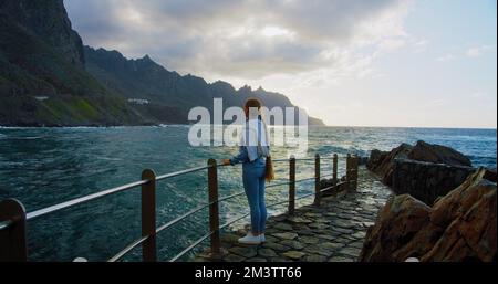 Frau genießt den Blick auf den stürmischen Ozean bei Sonnenuntergang. Vulkanische Berge von Anaga, Kanarische Insel, nördlich von Teneriffa. Wunderschöne Landschaften am Wasser. Stockfoto