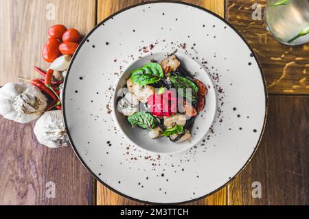 Blick von oben auf die Nudelgerichte. Schwarze Tagliolini-Pasta mit Garnelen auf weißem Teller. Knoblauch und heiße Paprika zur Dekoration. Hochwertiges Foto Stockfoto