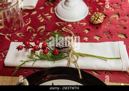 Festlicher Weihnachtstisch. Auf dem roten Läufer liegt eine weiße Serviette, dekoriert mit einem Holly-Zweig, mit einem schneebedeckten Zapfen, gebunden mit einer Schnur Stockfoto