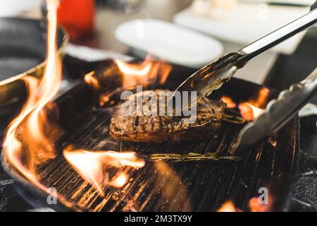 Bratverfahren für Striploin-Steak. Ein mittelgroßes, seltenes Stück Fleisch auf einer brennenden Grillpfanne, das mit einer Metallzange umgedreht wird. Hochwertiges Foto Stockfoto