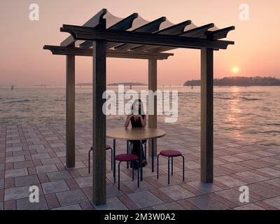 Junge weiße Frau, die auf einem Stuhl sitzt, mit einem runden Tisch davor unter einer einfachen Pergola an der Küste, mit einem Sonnenuntergang im Hintergrund, 3D Stockfoto